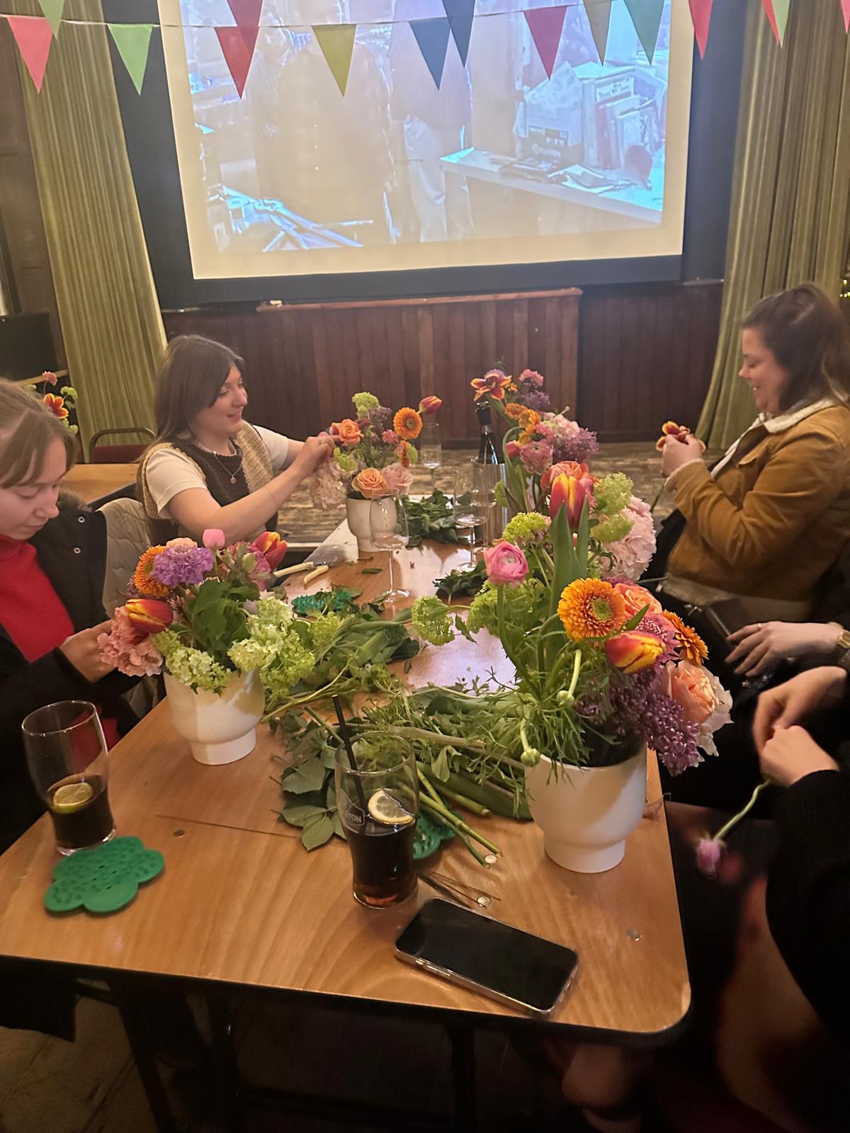 People sitting around a table with floral arrangements and drinks, with a screen displaying an image in the background.
