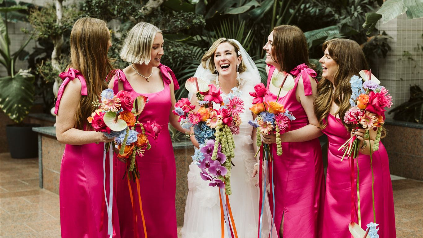 Bride and bridesmaids in pink dresses with colorful bouquets in an indoor garden setting.